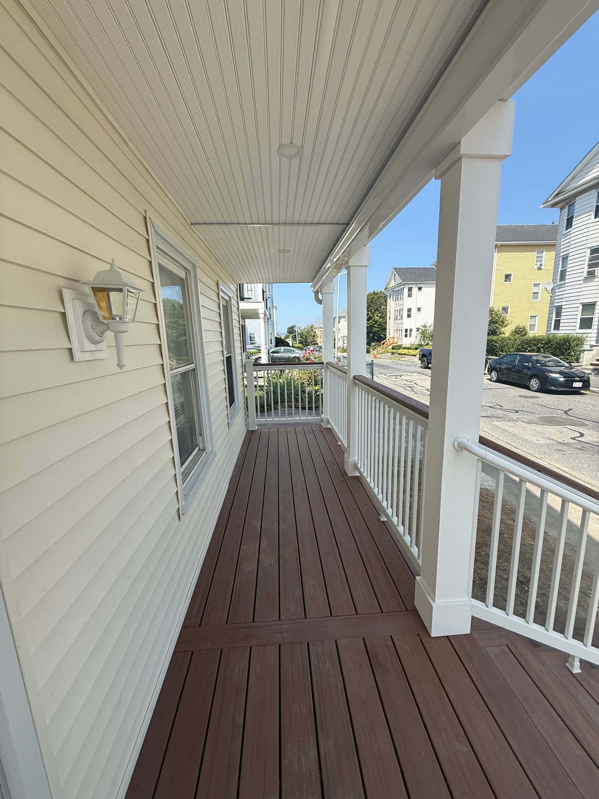 Covered porch with white columns, railing, and wood-toned deck. Siding is yellow, building in background.