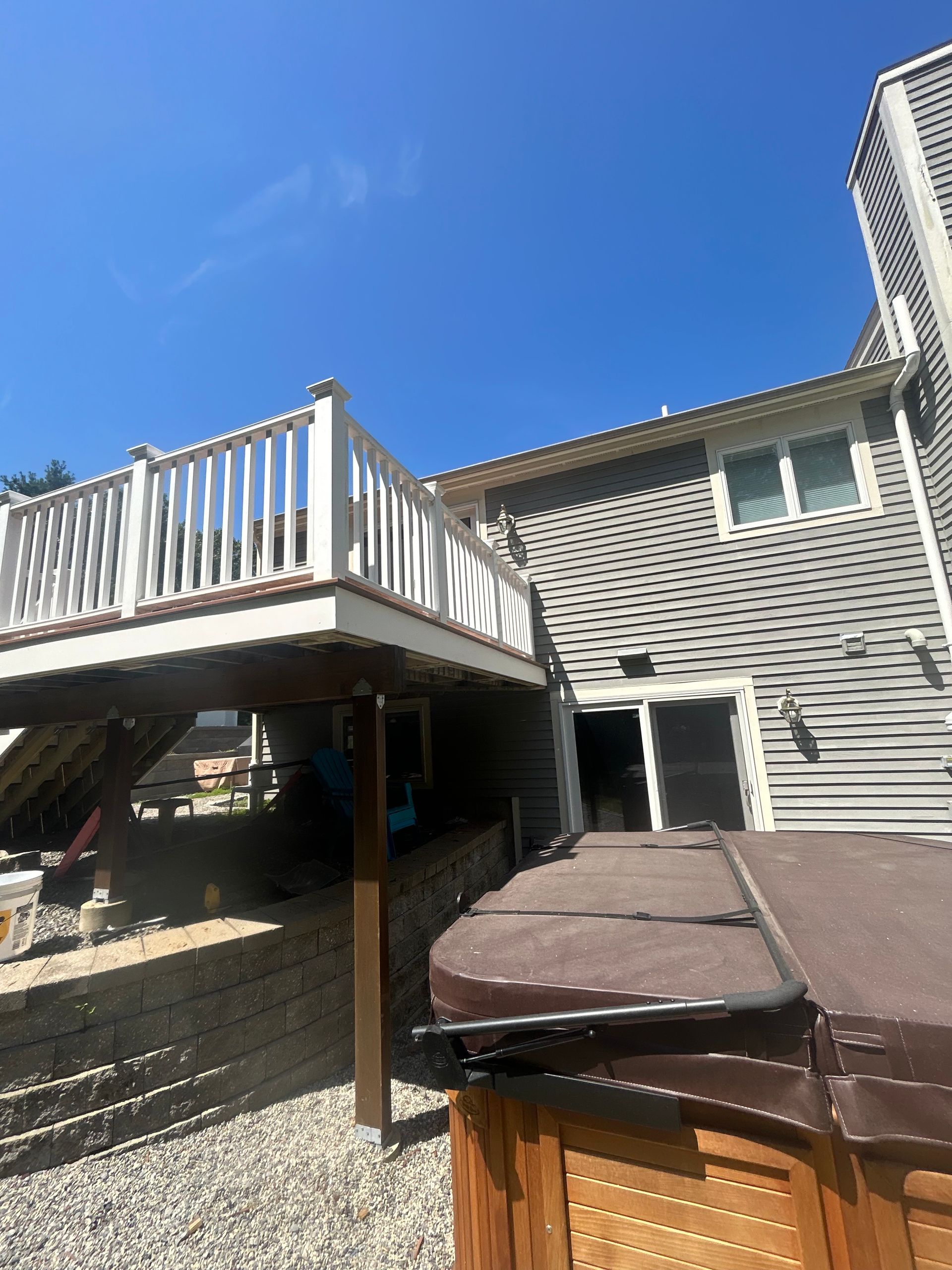 Elevated wooden deck with white railing, gray house, hot tub, and blue sky.