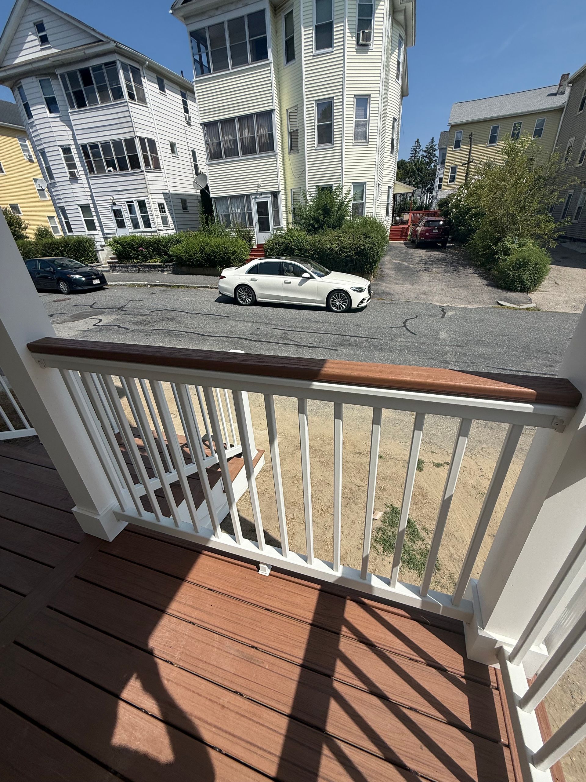 View from a deck with brown railings. White car parked in front of apartment buildings. Bright sunny day.