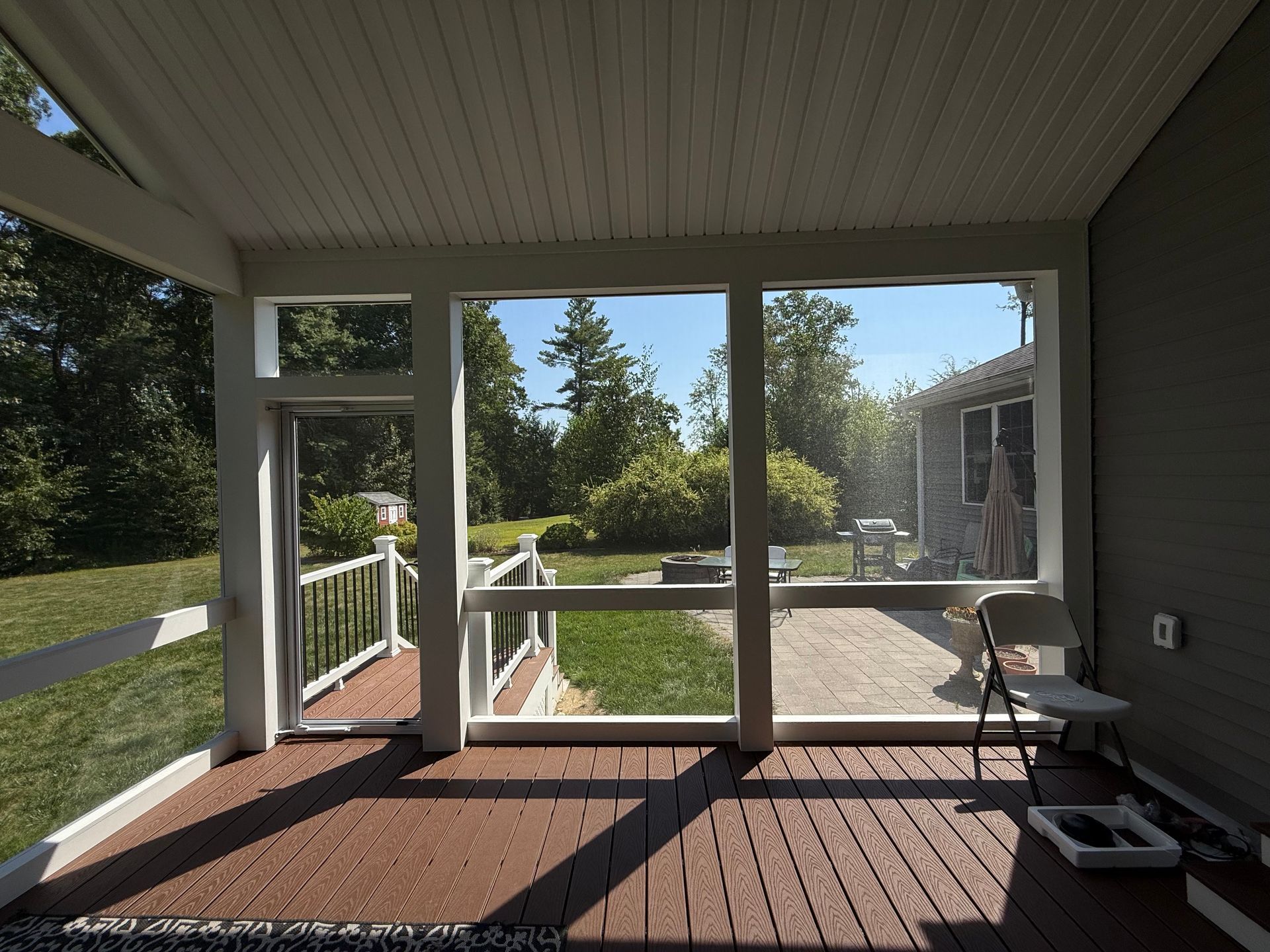 Screened porch with wooden deck, overlooking a sunny backyard with trees and a house.