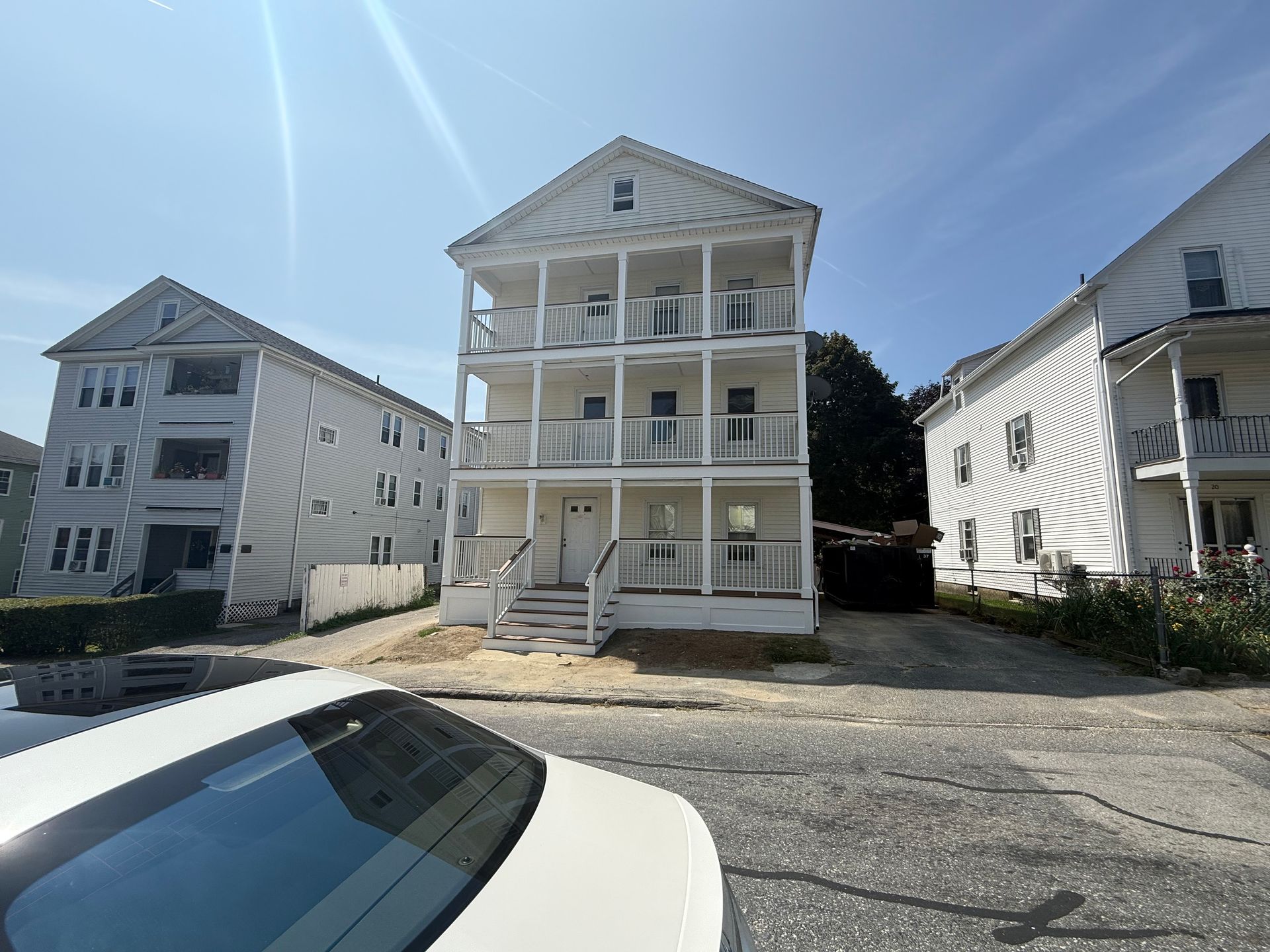 Three-story white house with balconies between similar houses on a sunny day; a car is in the foreground.