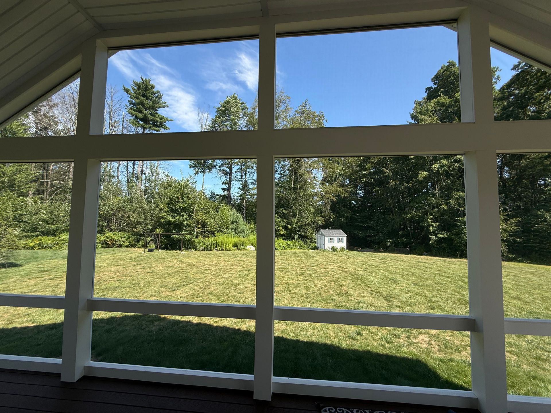 View from a screened porch, looking out at a grassy yard, trees, and a small white shed under a blue sky.