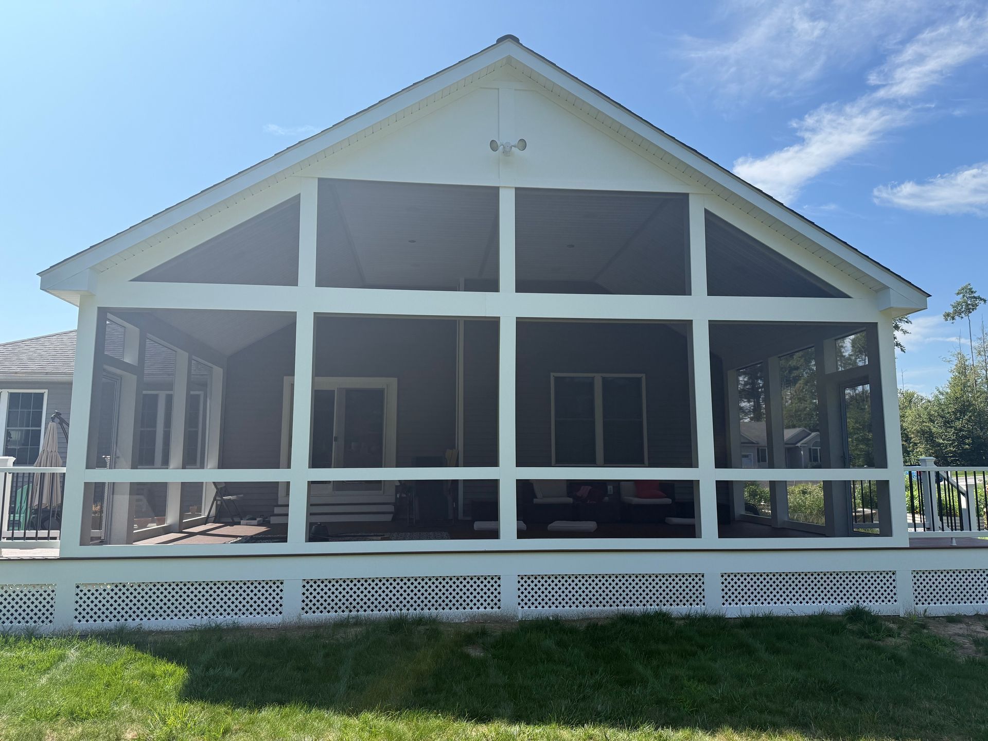 White screened-in porch with a gable roof and latticework base, attached to a house, with a blue sky.