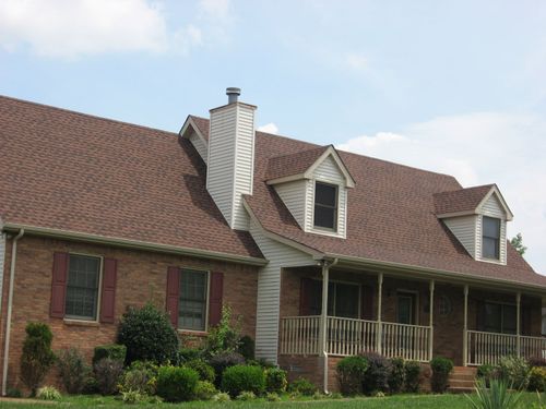 Brick house with a brown shingled roof, front porch, and trimmed shrubs under a blue sky