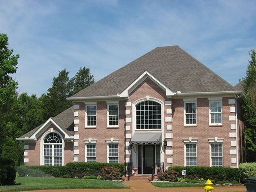Two-story red brick house with white trim, arched windows, and a manicured lawn under a blue sky