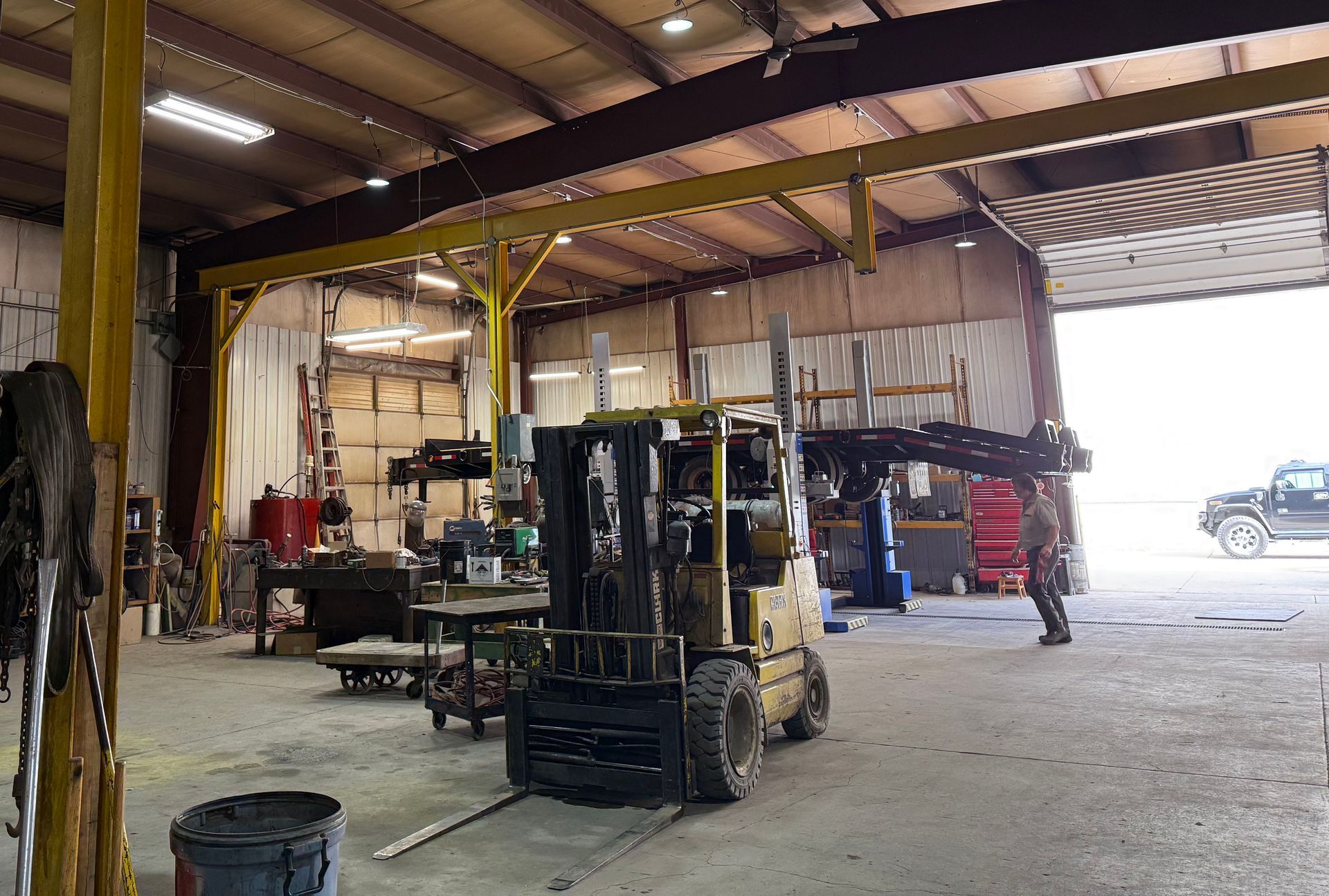 Yellow forklift in a workshop, moving metal. Man and truck in the background.
