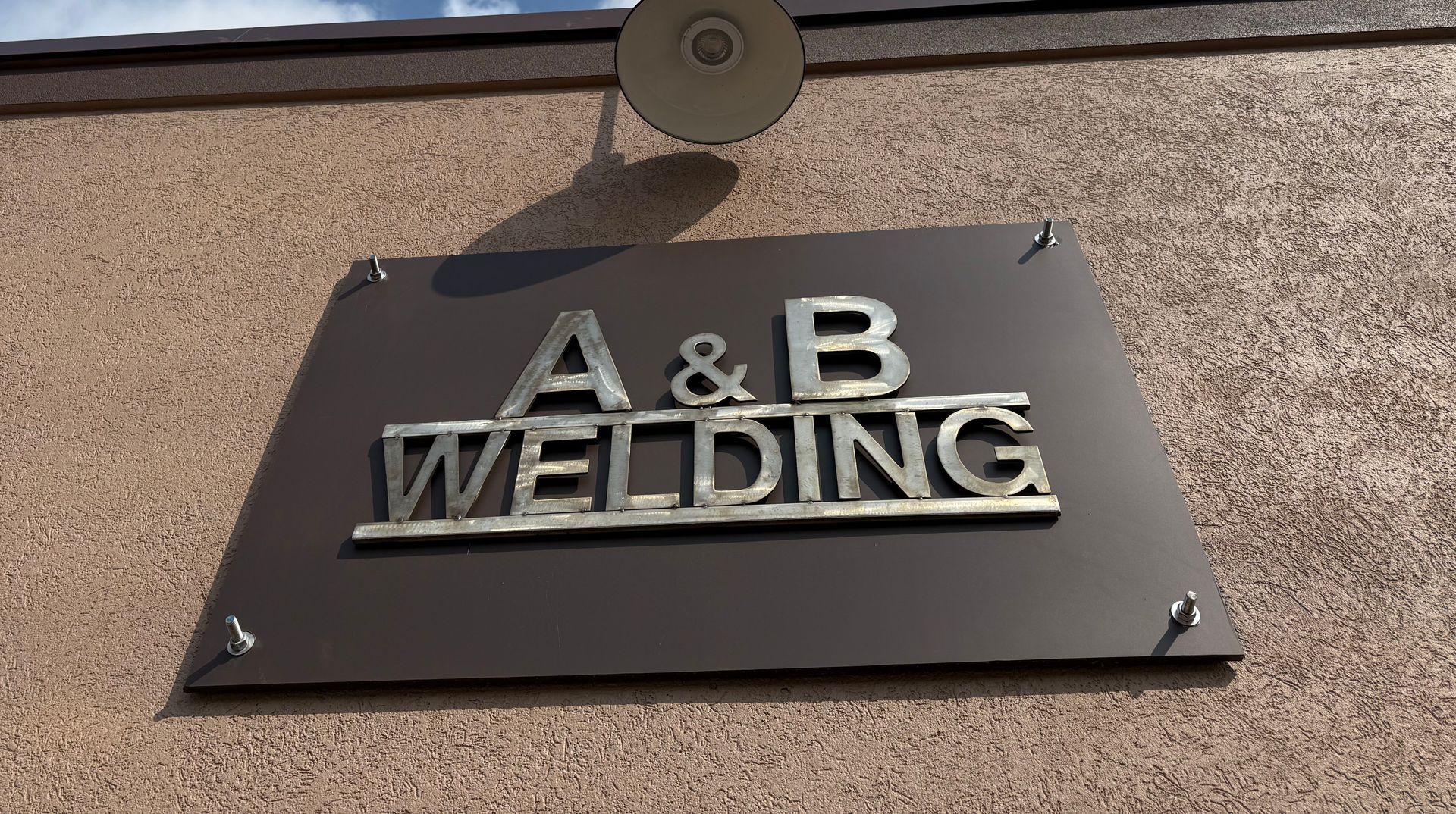 Sign for A & B Welding; silver letters on a brown trapezoid-shaped plaque, mounted on a tan wall.