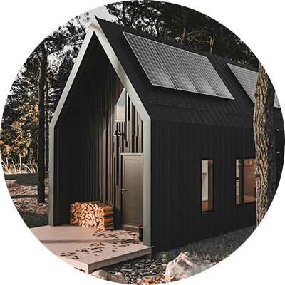 Black cabin in a forest with solar panels, a woodpile, and a wooden deck.