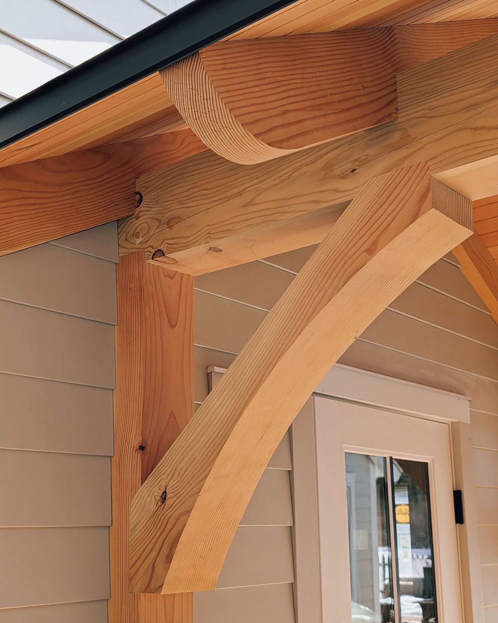 A close-up view of a decorative curved wooden bracket supporting the eave and beam of a building with light colored siding.