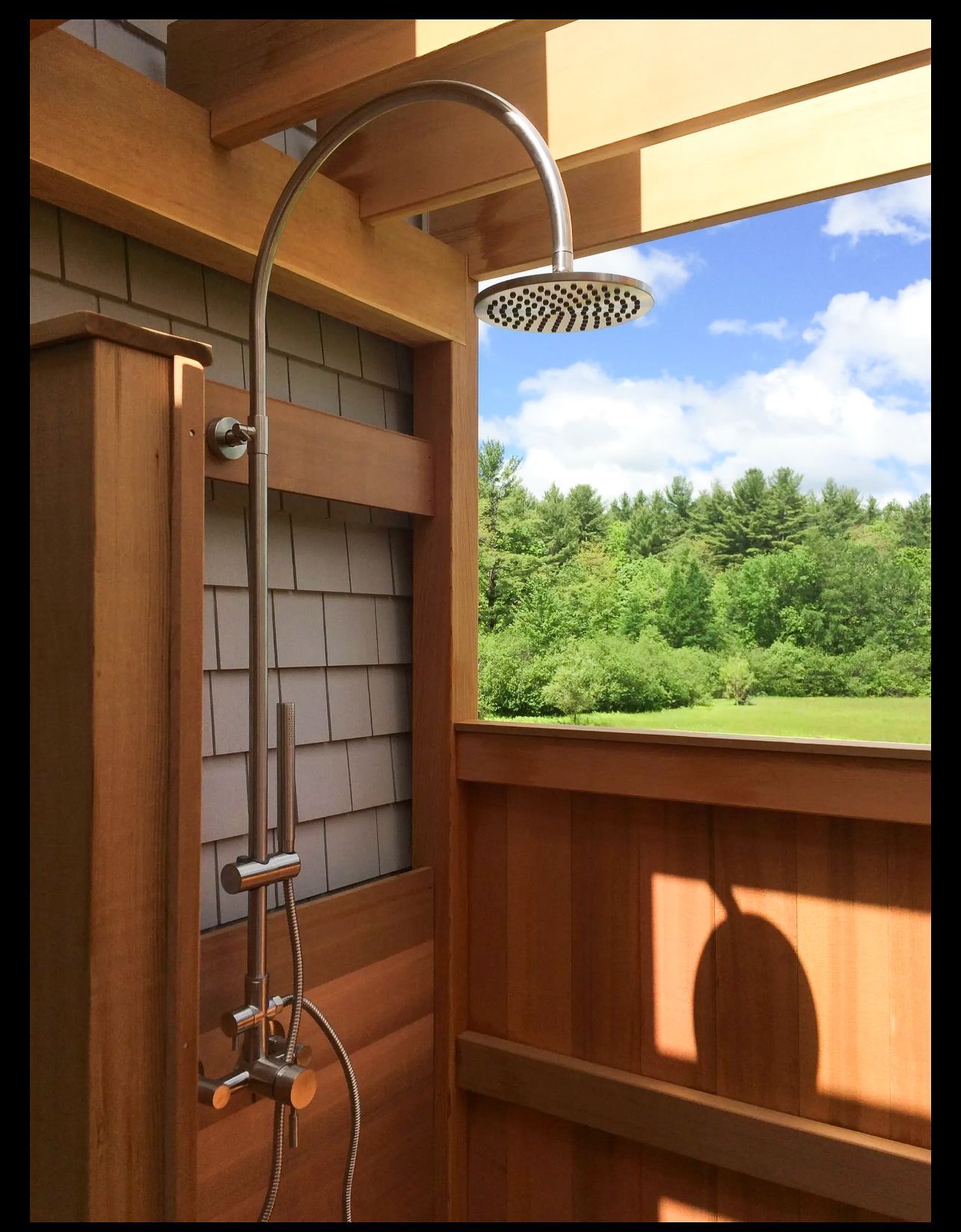An outdoor shower with a stainless steel rain showerhead and handheld attachment mounted on cedar wood walls.