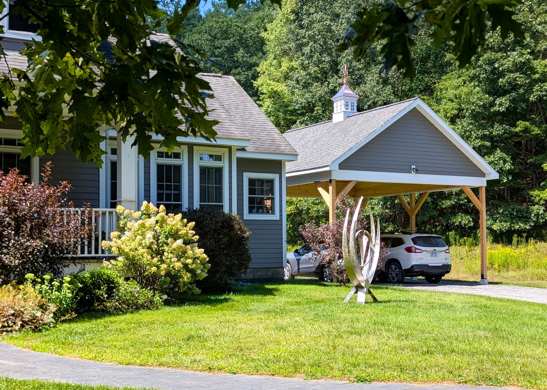 A gray house with a carport, a white SUV parked underneath, and a sculpture on the lawn.