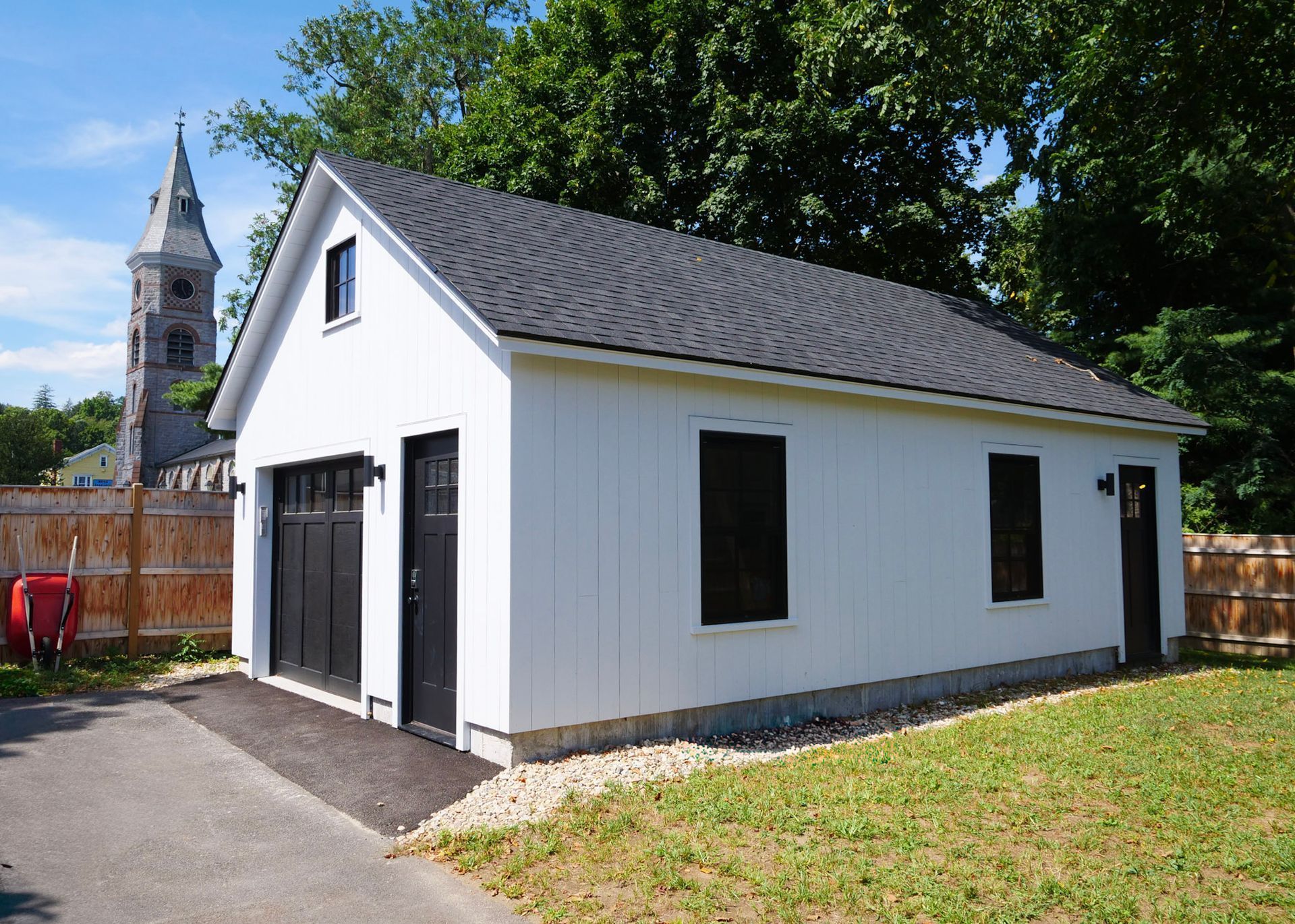 White garage with black doors and roof, asphalt driveway, church steeple in the background.