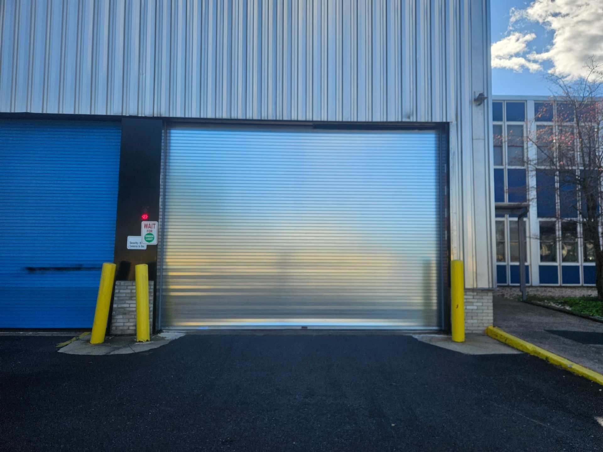 Metal roll-up garage door on a building, blue door to the left, yellow bollards.