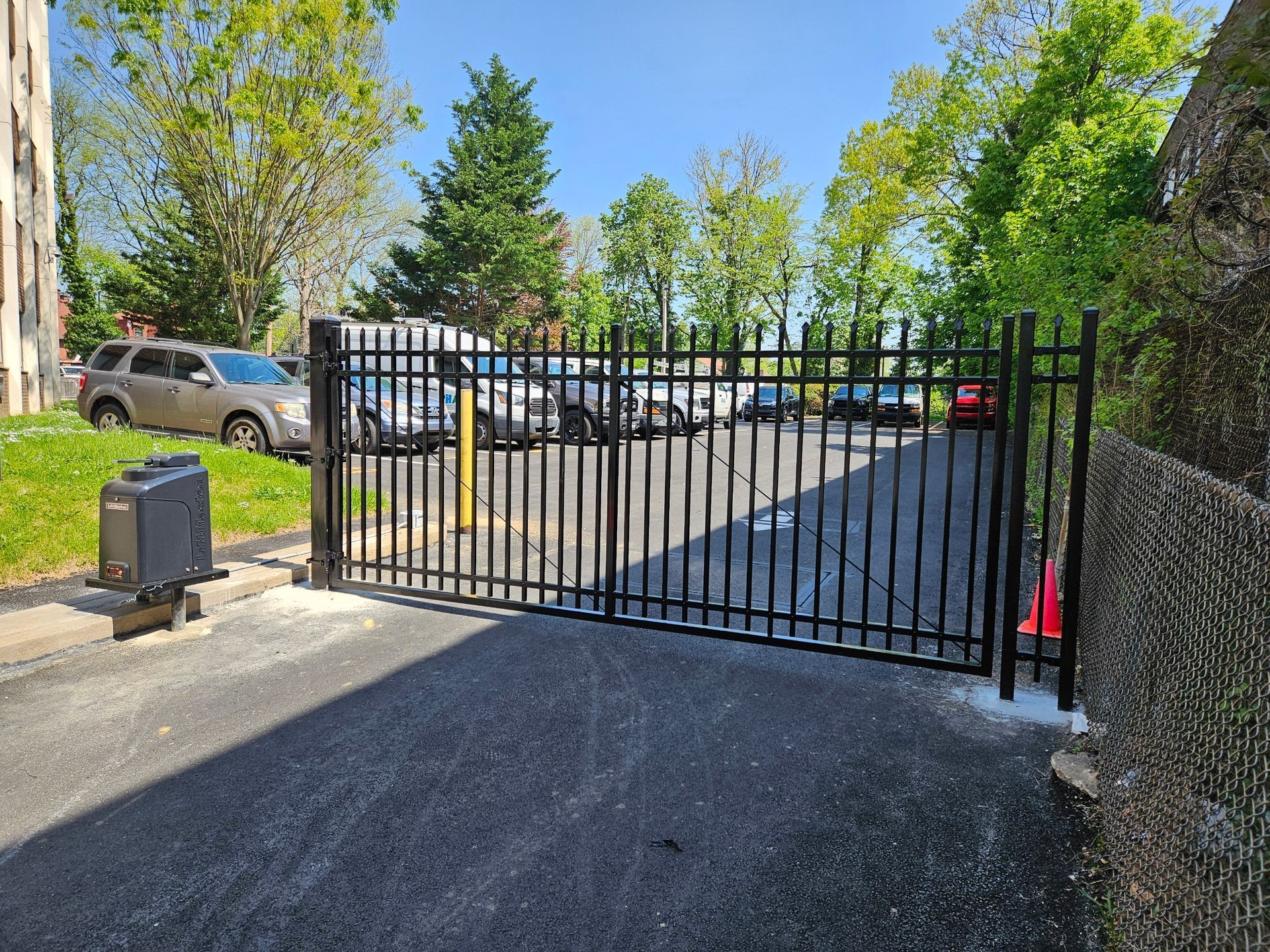 A black metal security gate bars a paved parking lot filled with various cars on a sunny day.