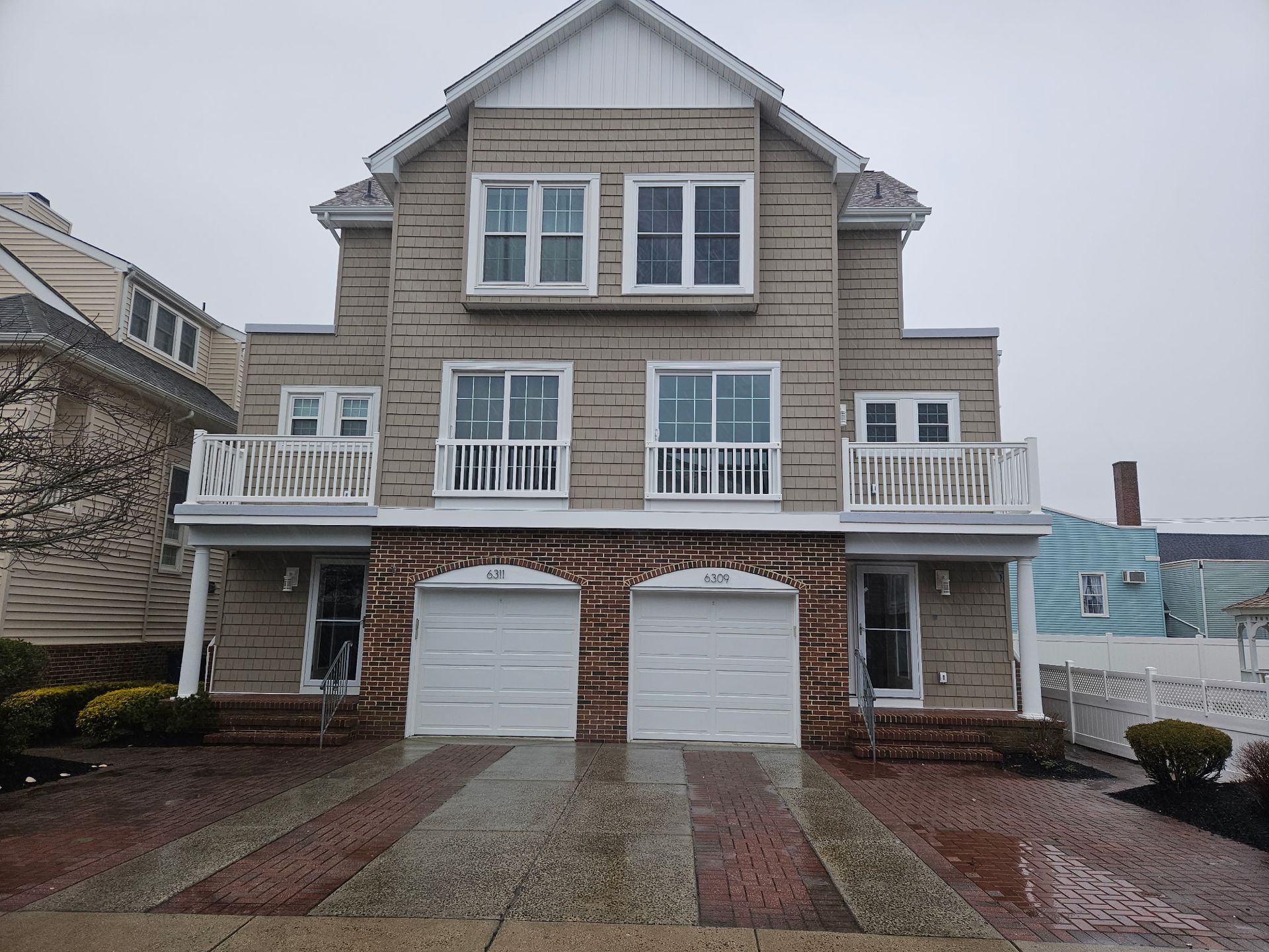 Two-story beige brick house with white balconies and garage doors; wet brick driveway.