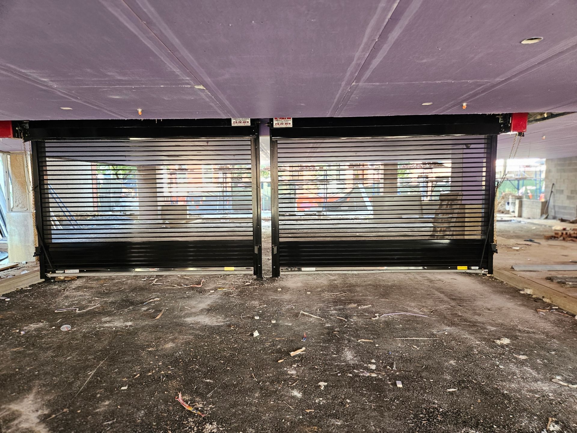 Two black metal rolling security gates are closed side-by-side in a dim, unfinished parking area.