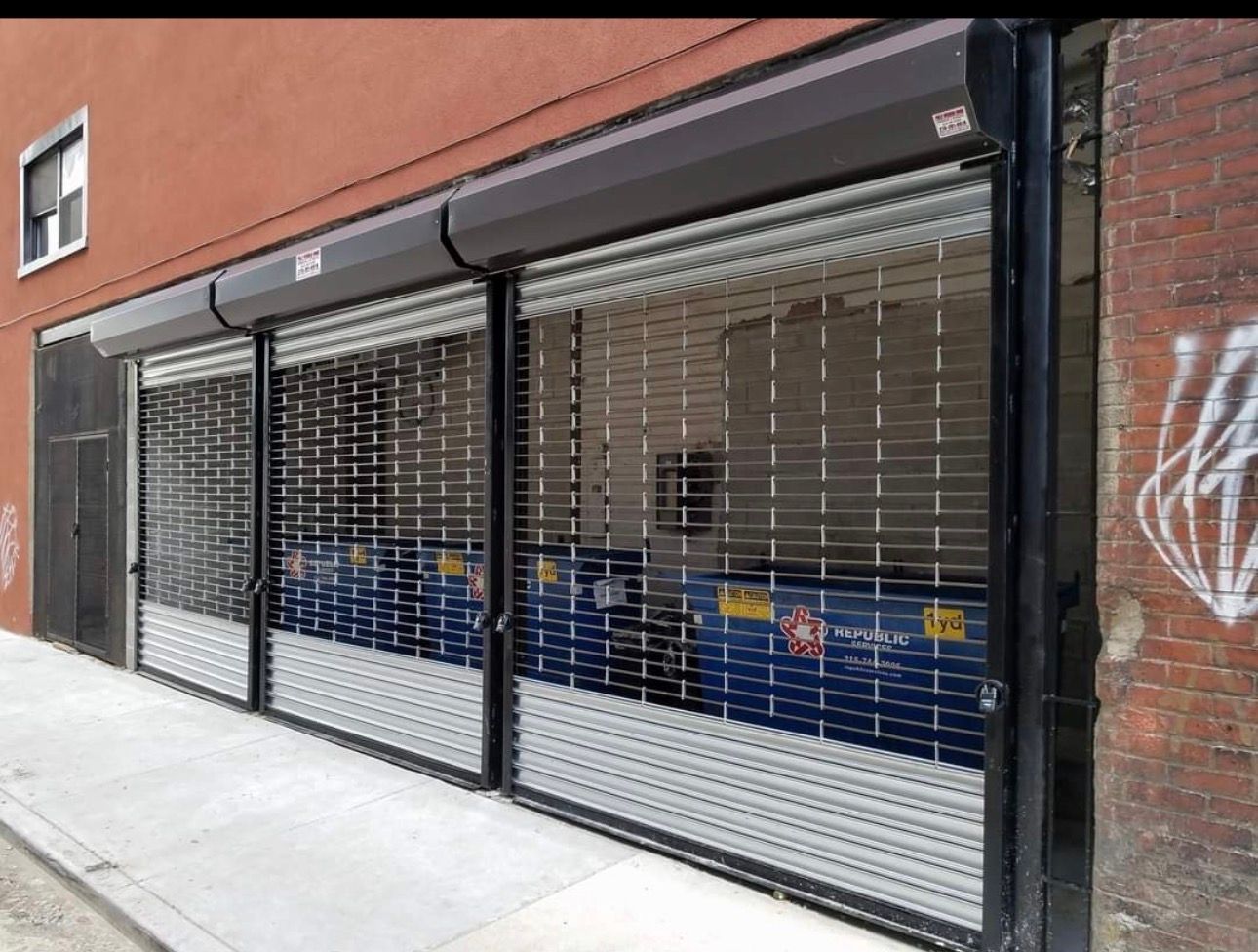 Three black metal rolling security grilles covering an alley entrance with two large blue dumpsters visible inside.