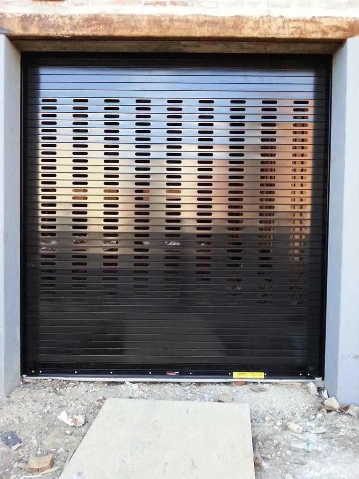 Black rolling shutter door in a gray doorway, with horizontal rectangular openings. Concrete and debris below.
