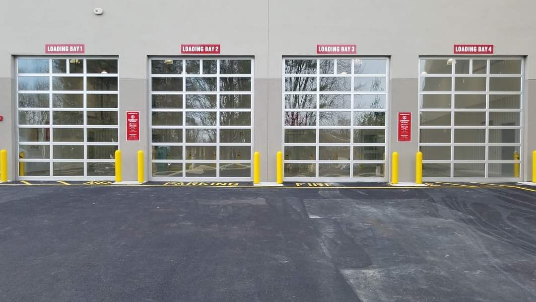 Four garage doors with clear panels, red signs above, yellow bollards.