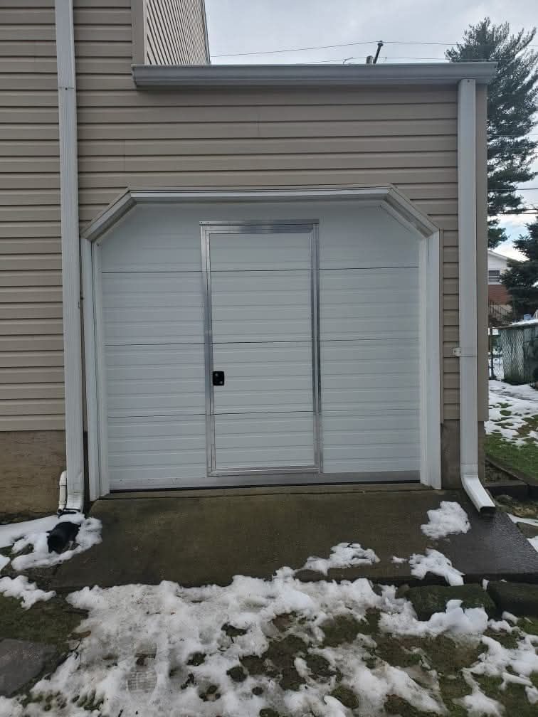 White garage door with an inset door, exterior siding, and snow on the ground.
