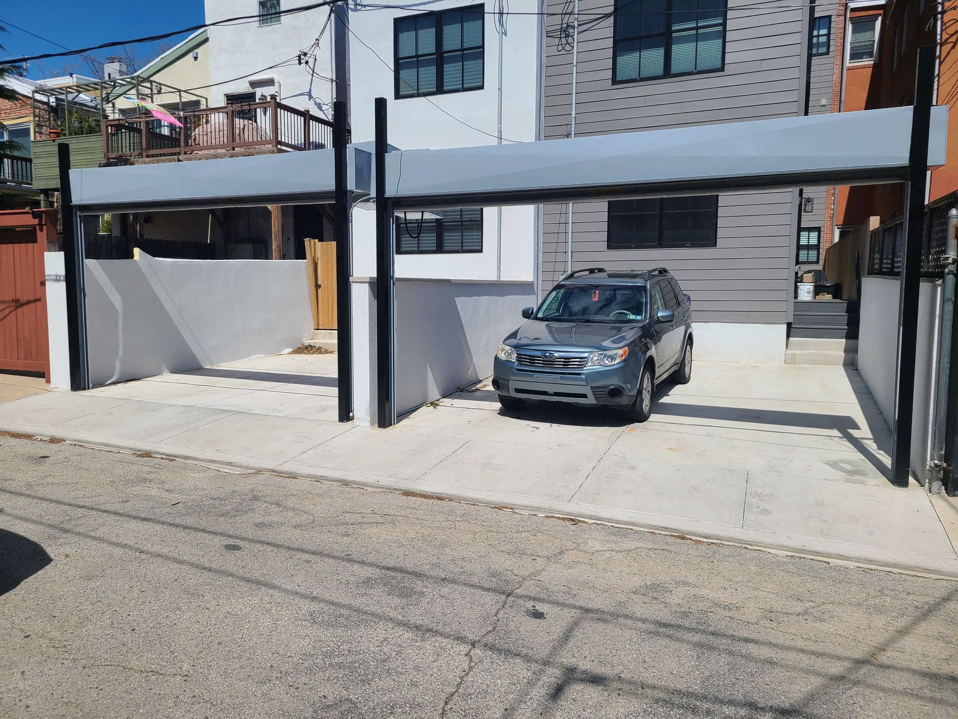A gray SUV parked in a driveway under a metal awning. Buildings flank the space.