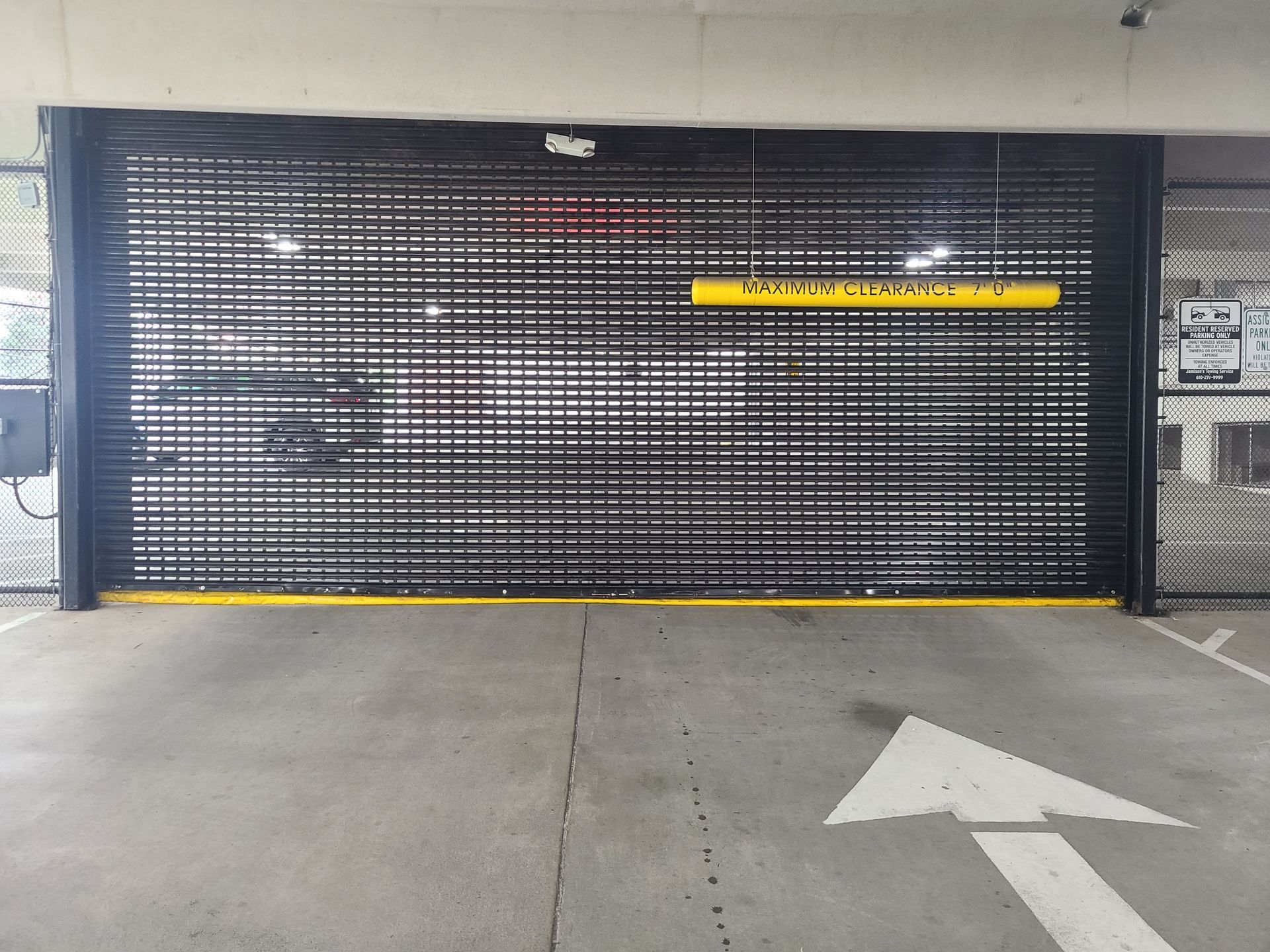 Roll-up gate at a parking garage entrance. Yellow height restriction bar. White arrow on concrete floor.