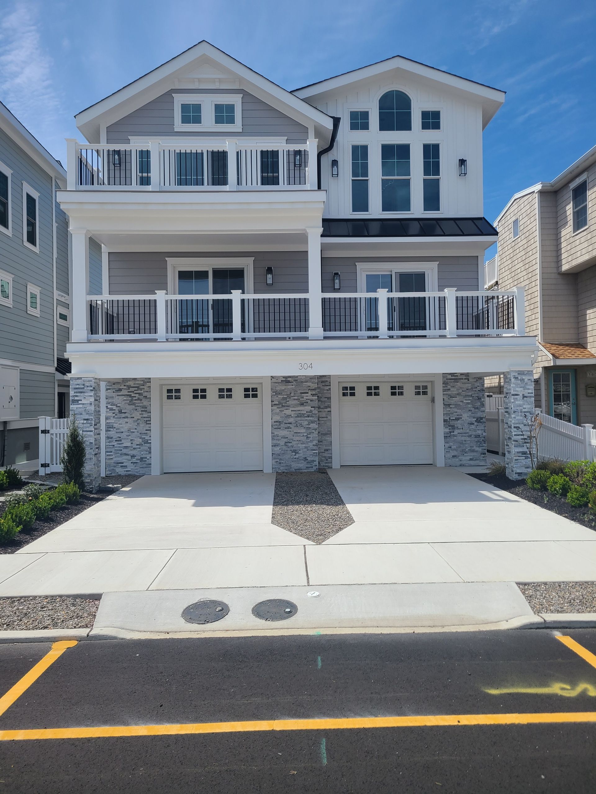 Two-story beach house with light gray siding, white trim, balconies, two garage doors, and blue sky.