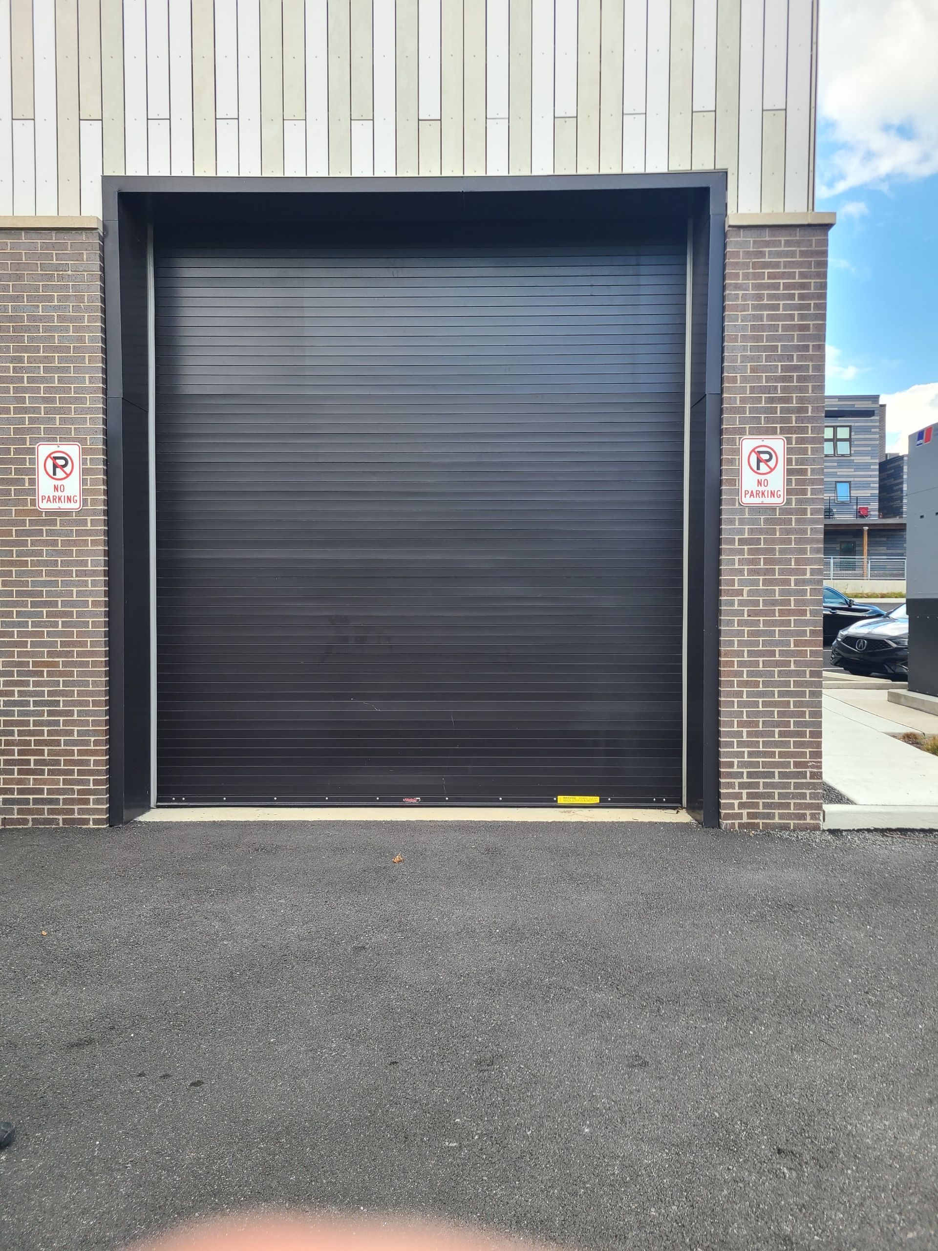 Black garage door framed by brick and a light-colored building with warning signs on either side.