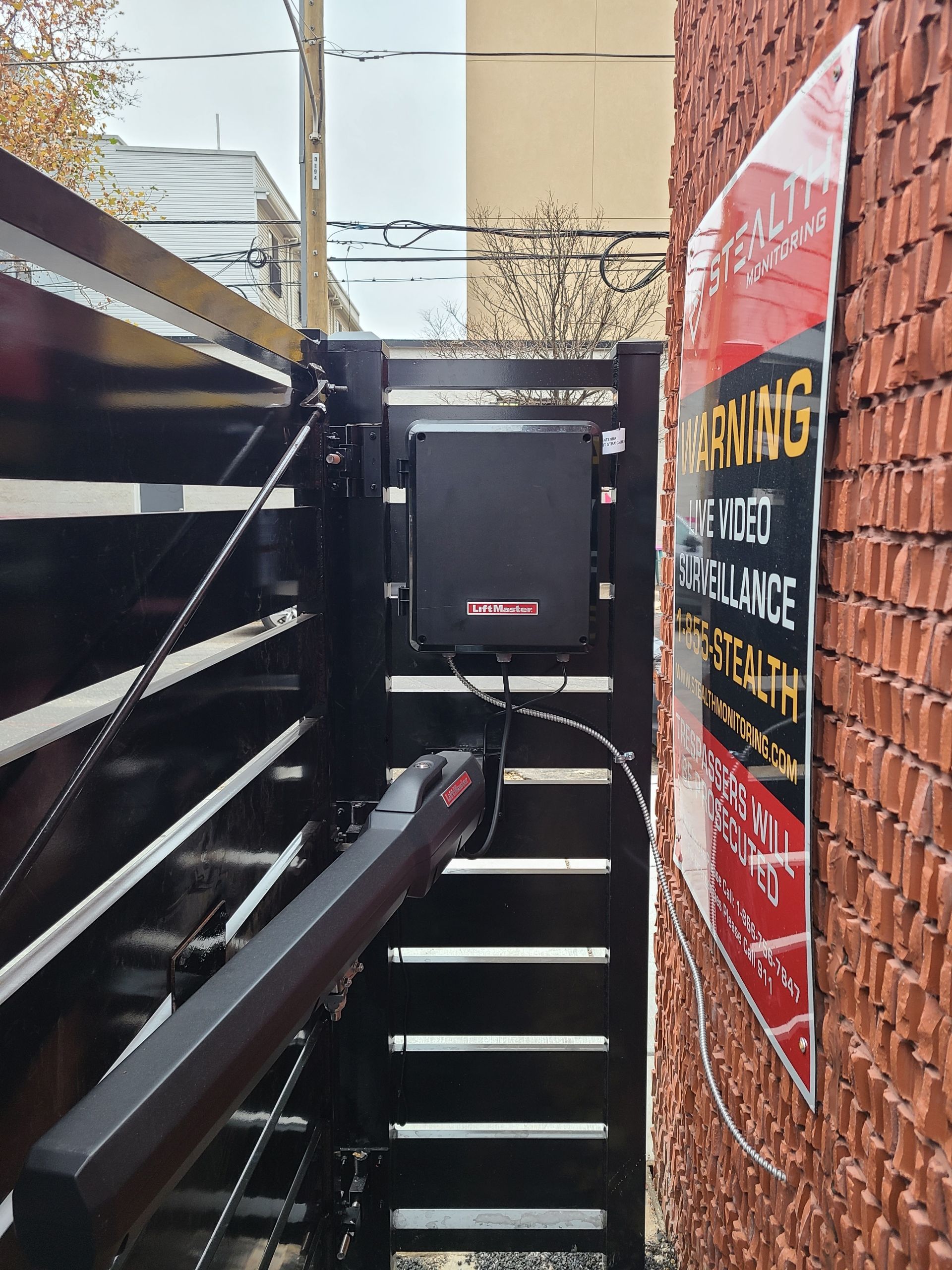 Black automatic gate with a warning sign on a brick wall.