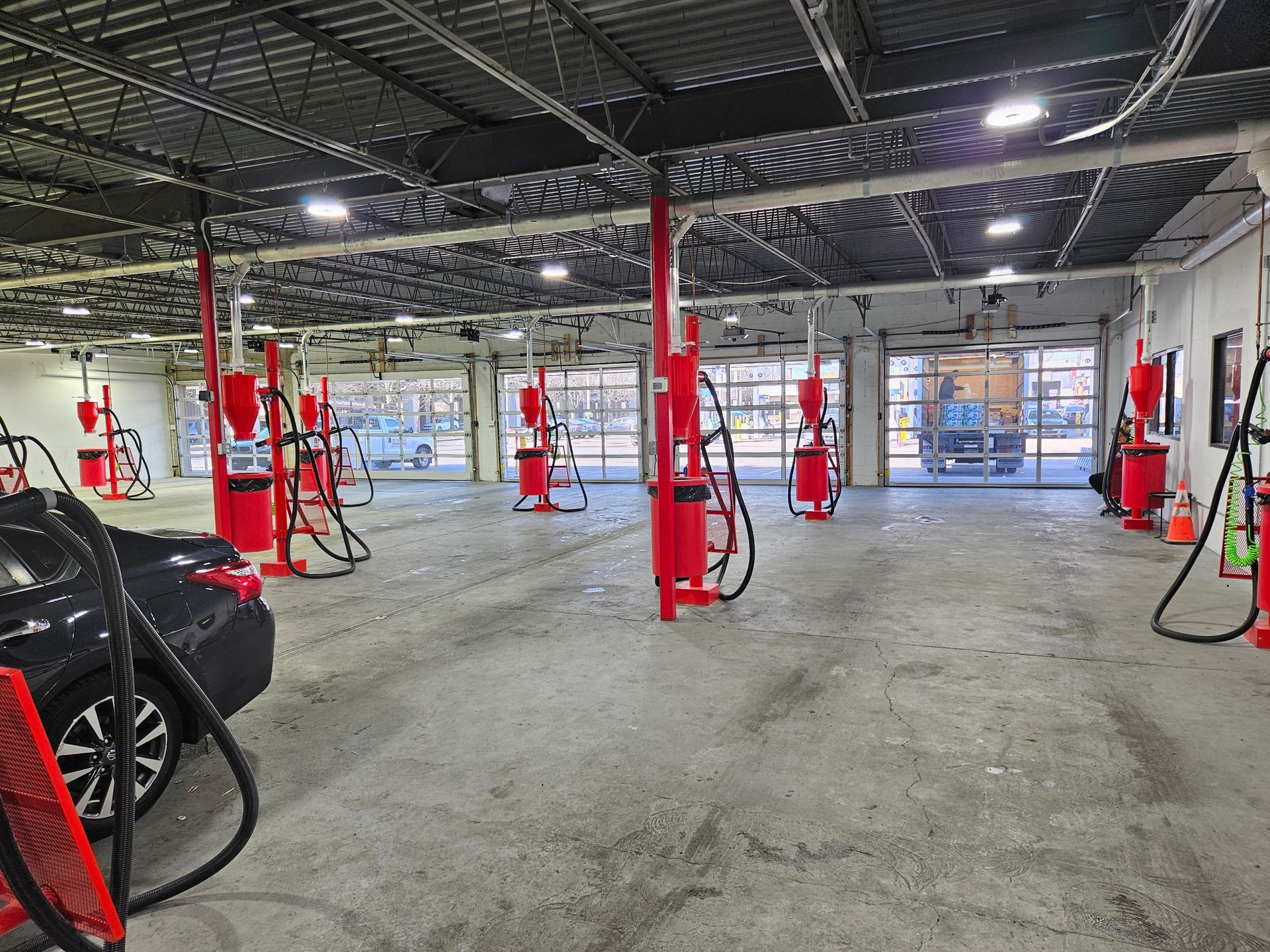 Interior of a car wash. Red vacuum stations line the concrete floor. Open bay doors reveal outside light. A black car is visible.