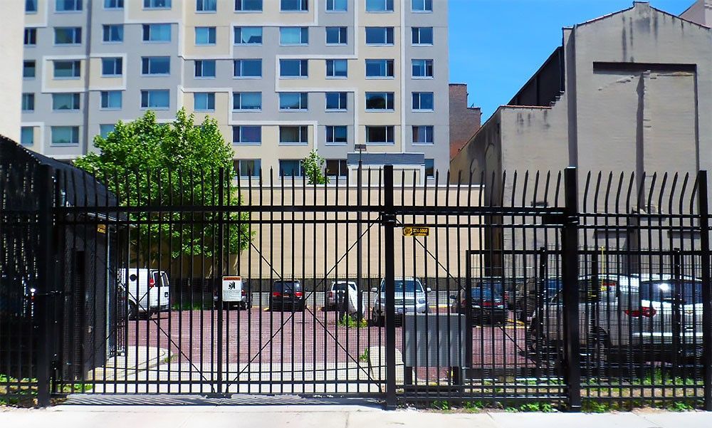 Black metal gate leads to a parking lot with cars, next to a tall building and a tan structure.