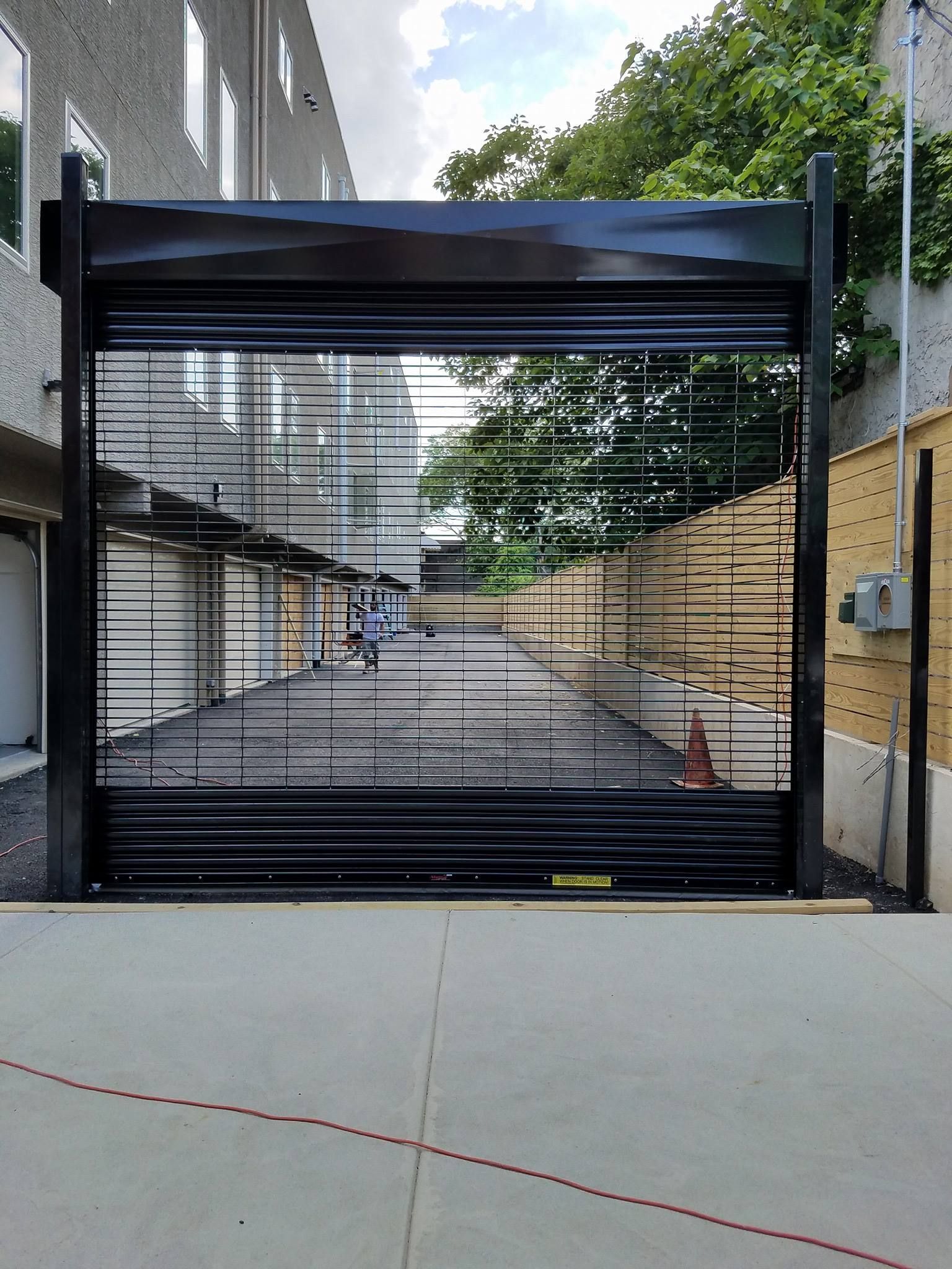 Black metal security gate on a concrete driveway. Slatted design, open, revealing a view of a street.