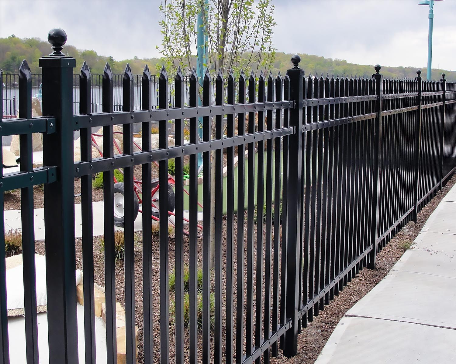 Black metal fence near a sidewalk, with pointed tops, protecting a landscaped area.