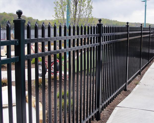 Black metal fence with vertical bars, along a sidewalk near foliage and water.