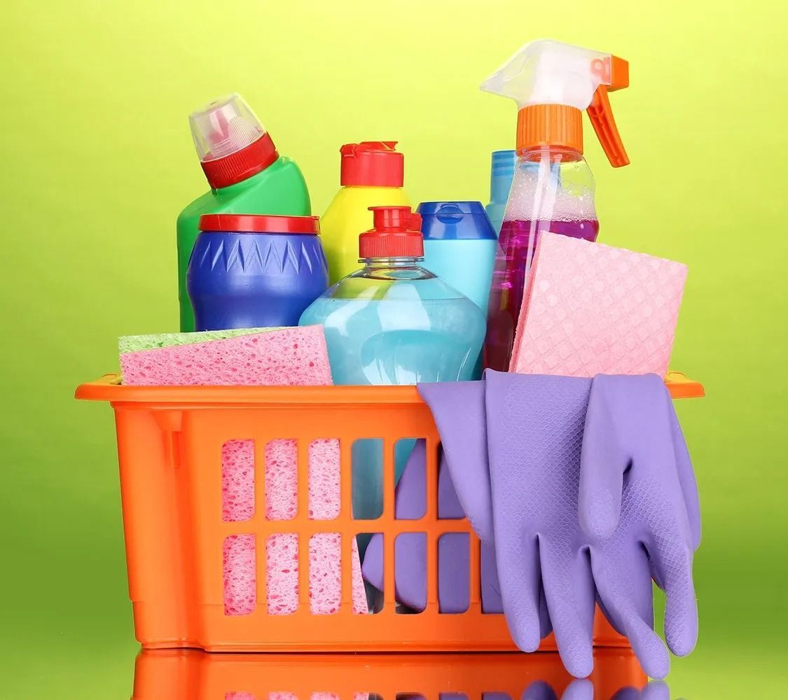 Orange cleaning basket with colorful spray bottles, sponges, and purple gloves on a green background