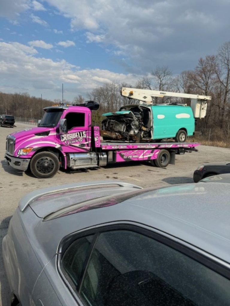 Pink tow truck carrying a teal lift boom on a roadside lot under a partly cloudy sky