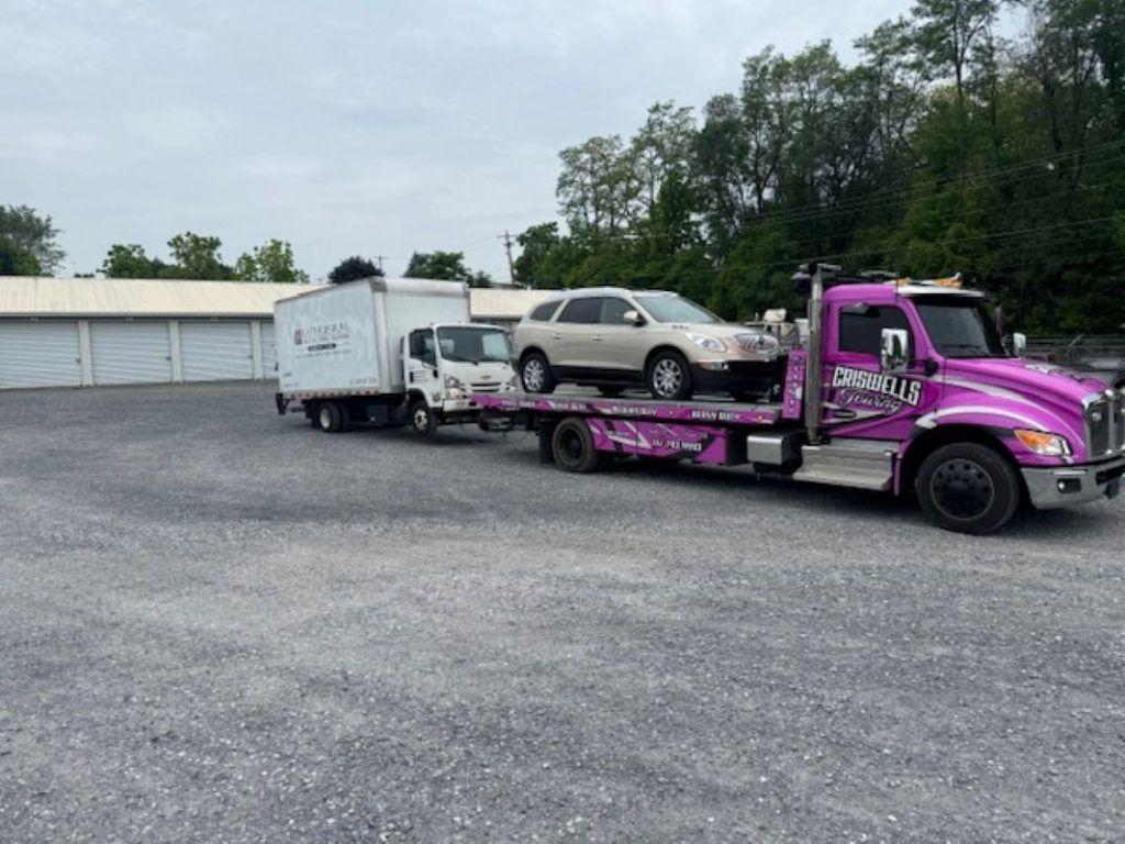 Magenta tow truck hauling a beige SUV in a gravel lot beside white buildings and trees