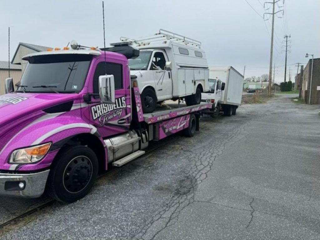 Purple tow truck hauling a white utility truck on a wet street under an overcast sky