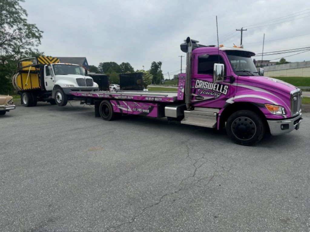 Purple flatbed tow truck hauling a white utility truck in a parking lot