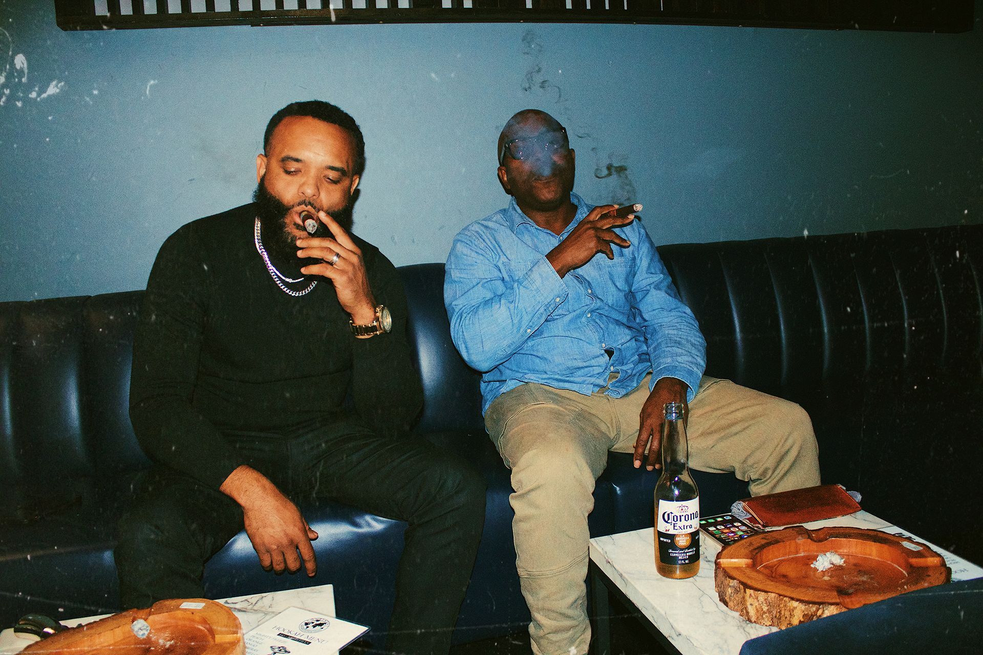 Two people sit on a black leather couch, smoking and drinking in a dim lounge with a pizza box on the table.