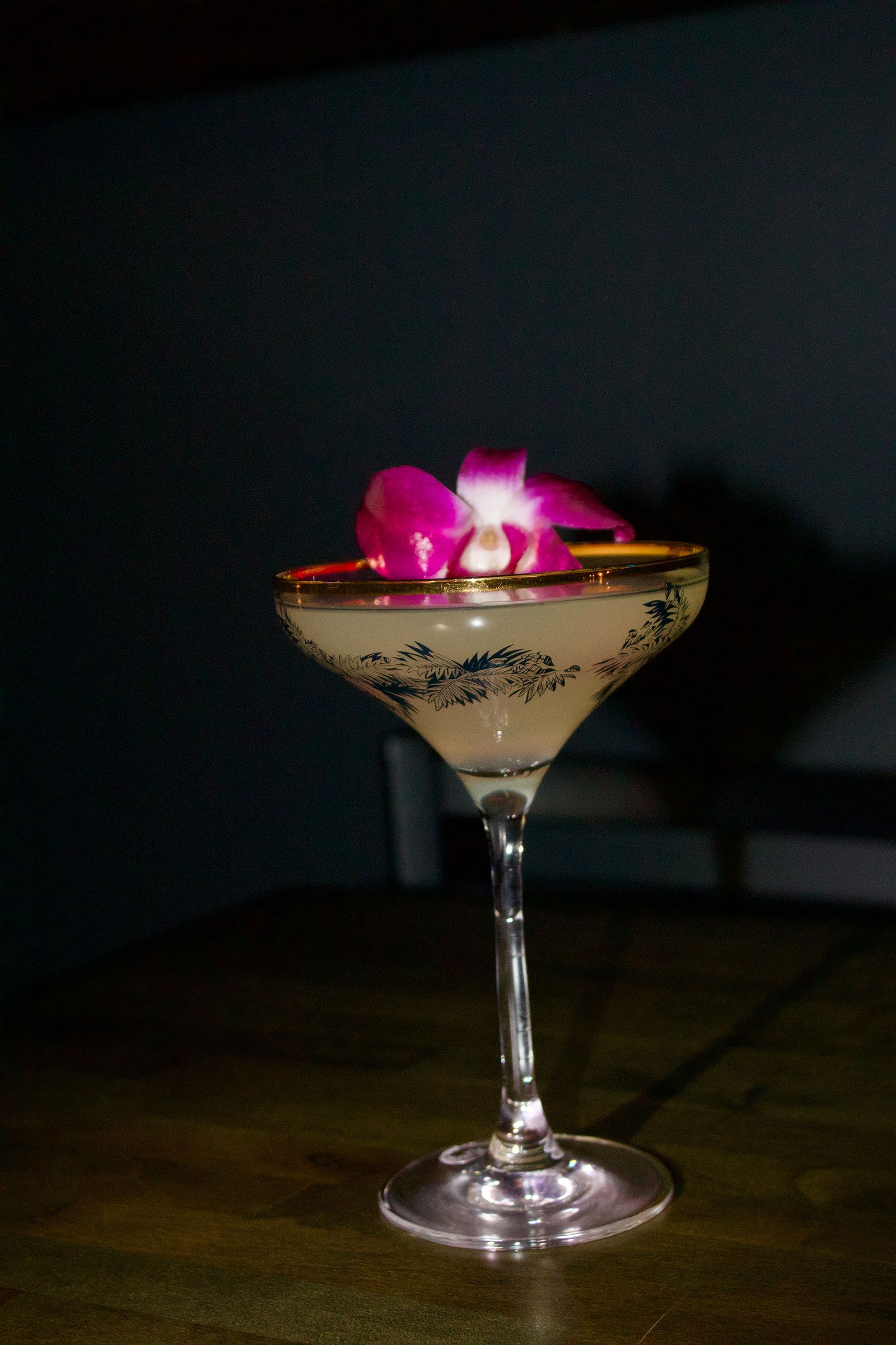Martini glass with a pink flower garnish on a dark tabletop against a black background