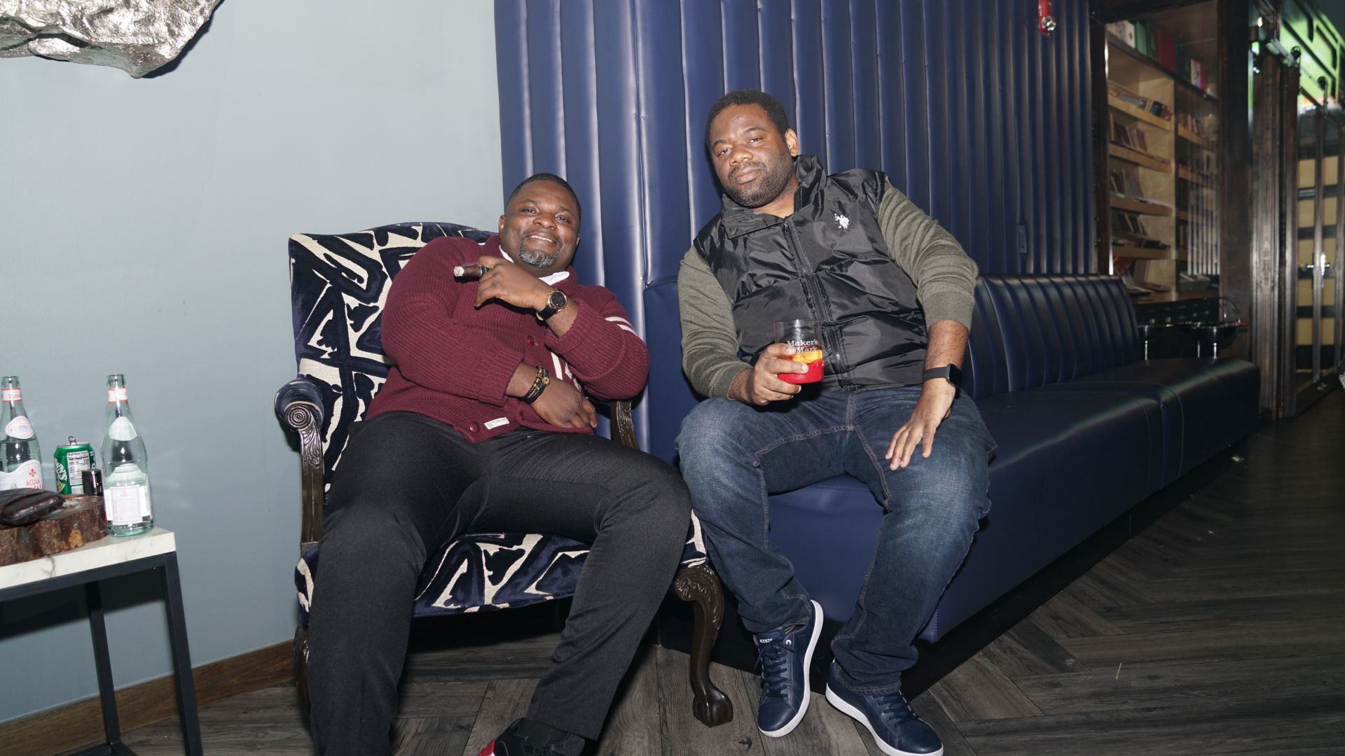 Two people sitting on a couch in a dimly lit lounge, smiling and holding drinks.