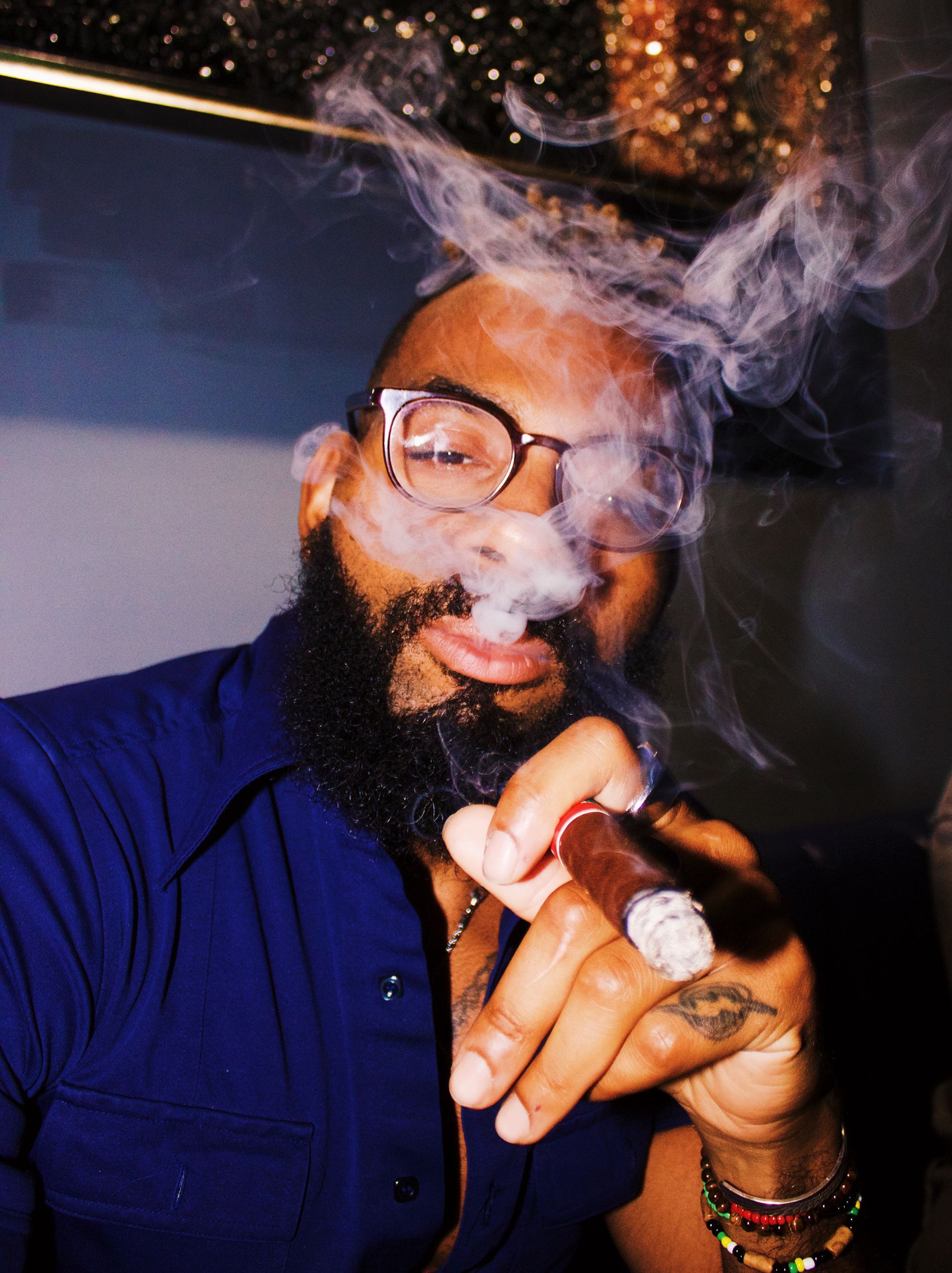 Person in glasses smoking a cigar, exhaling smoke in a dimly lit setting.