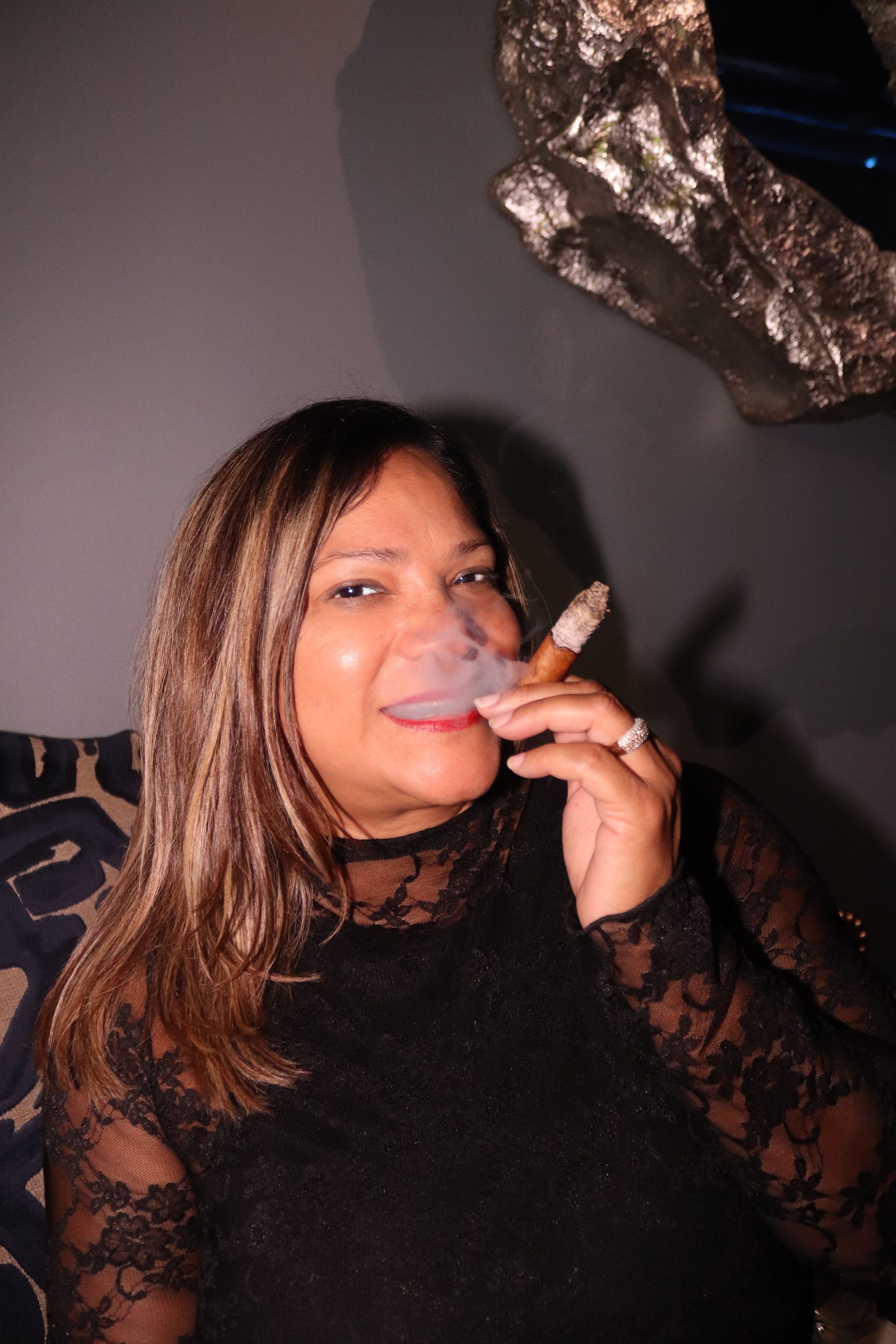 Woman in a black lace top smiling while holding a cigar, seated indoors against a dark wall.