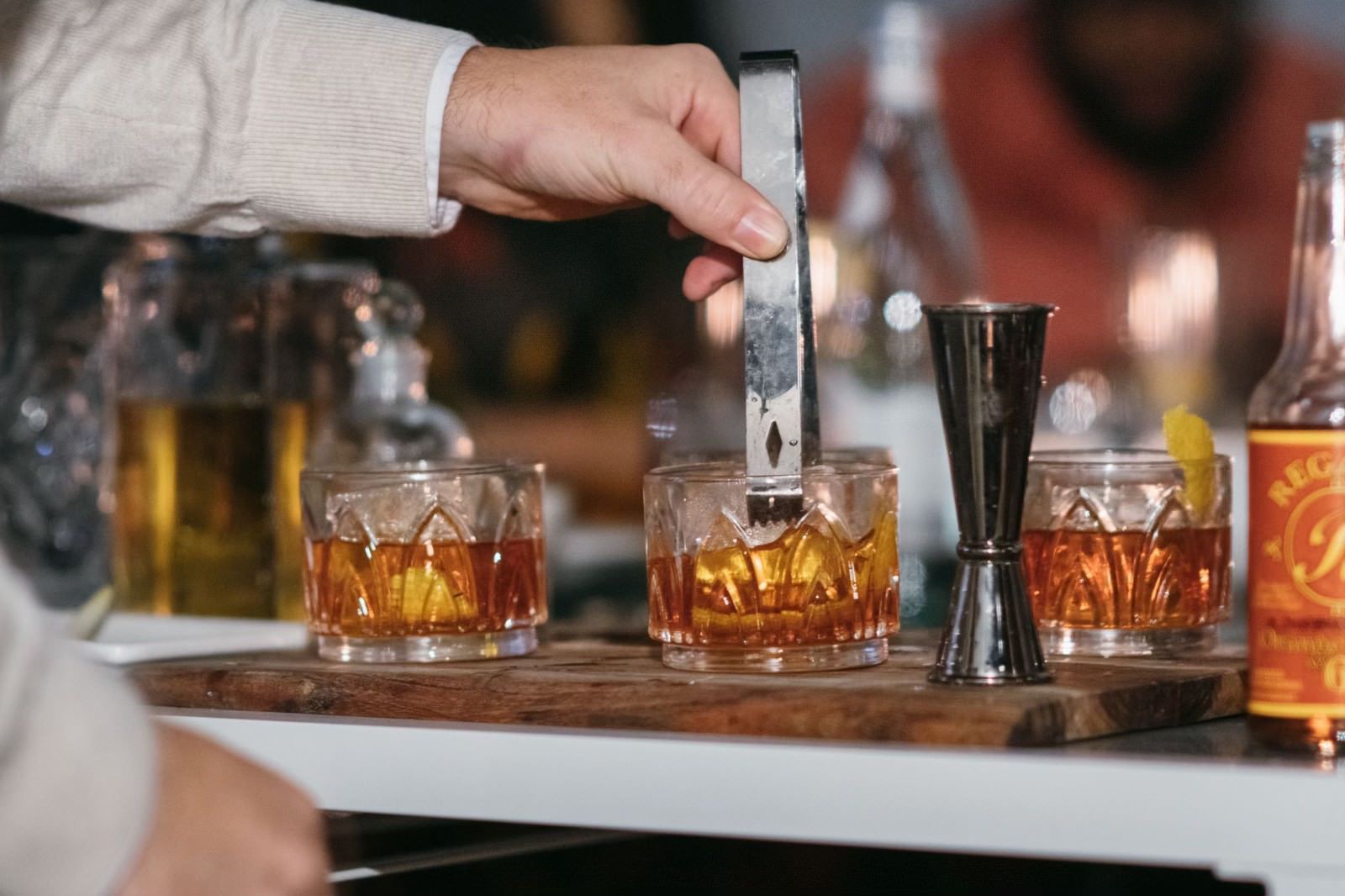 Hand pouring a drink into a rocks glass on a bar counter with a jigger nearby