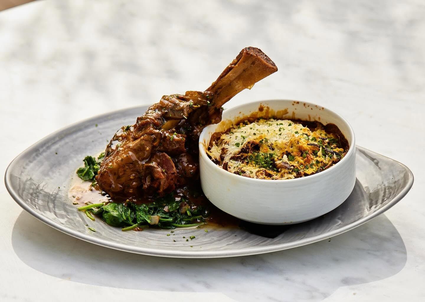 Braised lamb shank with herb garnish beside a bowl of creamy mashed potatoes on a gray plate