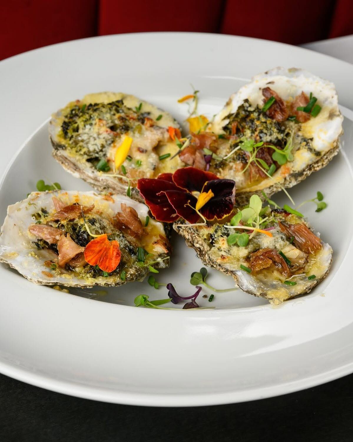 Four oyster shells topped with garnishes on a white plate, served on a red tablecloth