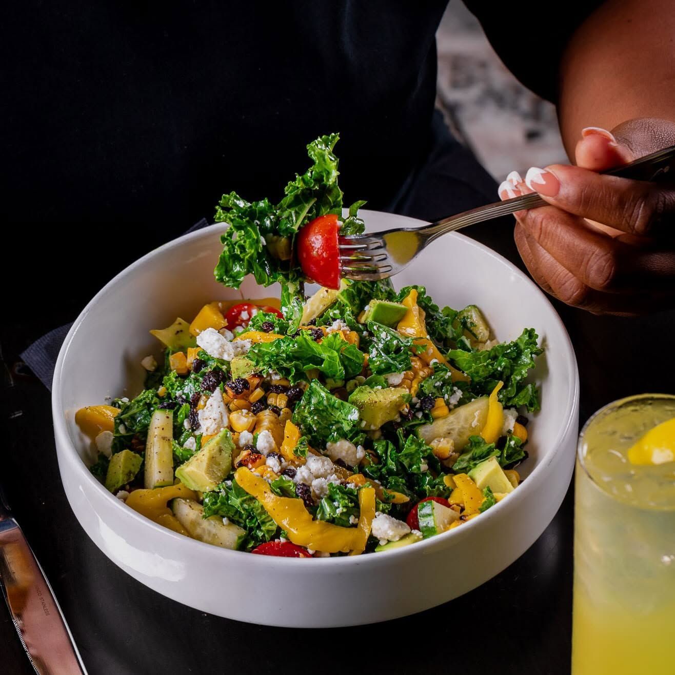 A bowl of colorful salad with kale, fruit, and a hand holding a fork; a drink sits beside it.
