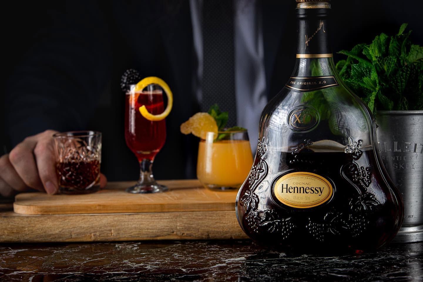 Hennessy bottle on a bar with cocktails, lemon garnish, and glasses in a dark lounge setting