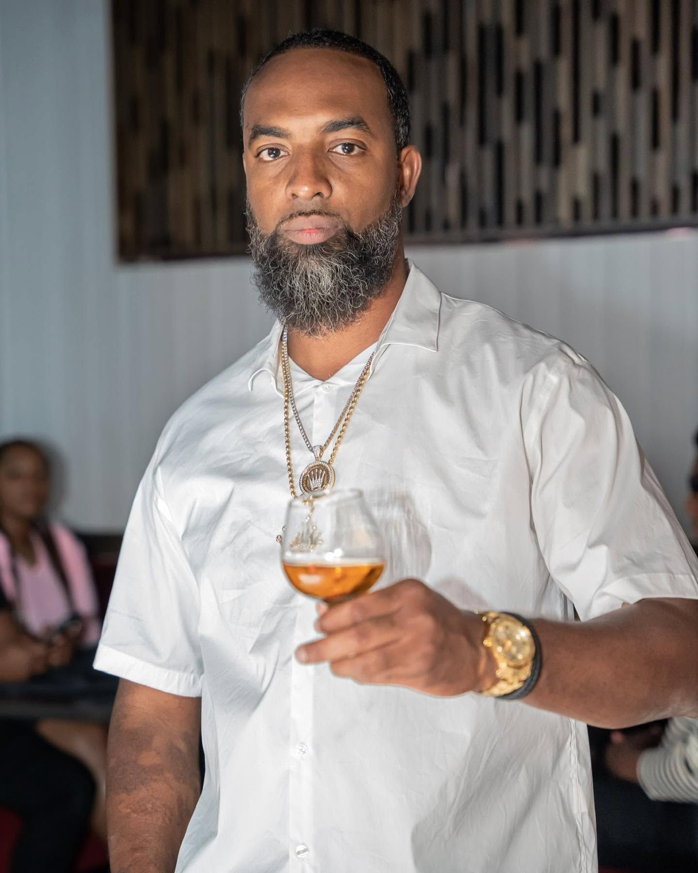 Man in white shirt holding a cocktail glass indoors, wearing gold jewelry.